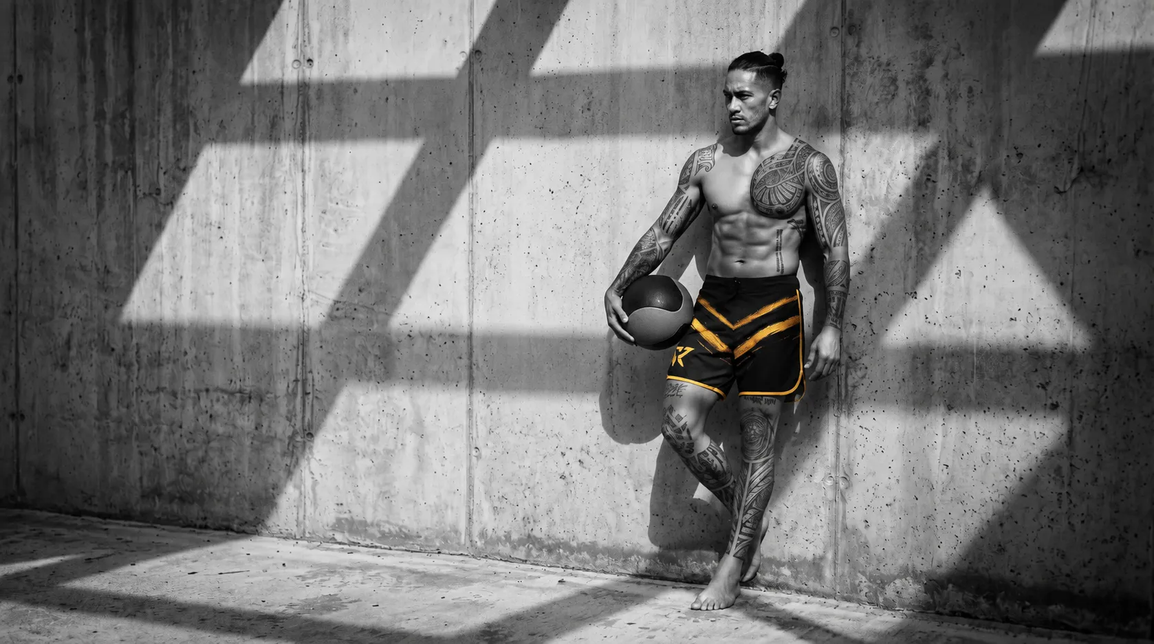 A muscular man with extensive tribal tattoos, wearing black and gold athletic shorts, leans against a textured concrete wall, holding a medicine ball. Dramatic light and shadow patterns fall across the scene.