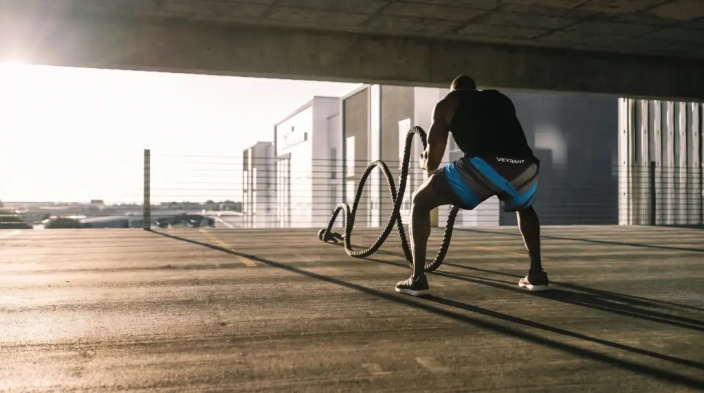 Man in athletic shorts and t-shirt performing battle rope workout in a sunlit parking garage, showcasing strength and fitness, wearing premium athletic shorts.