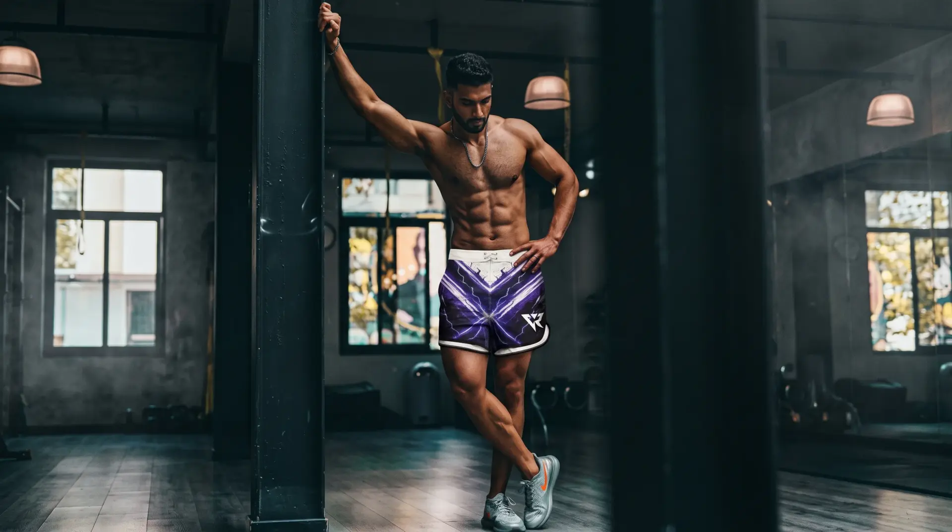 Fit man in purple lightning shorts leans against a gym pillar, showing off abs and muscular physique and showcasing Veyrant "Rift" competition shorts.