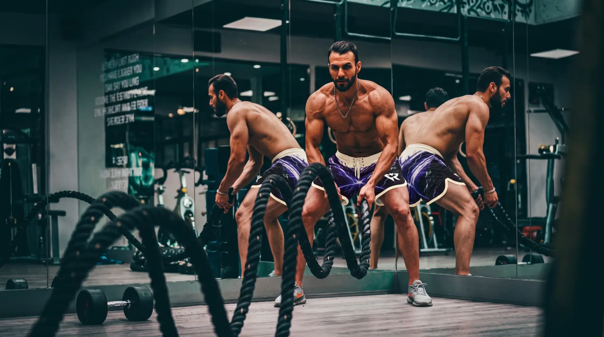 Muscular man with beard and wearing premium men's physique competition shorts performing battle rope exercise in a gym, reflected multiple times in mirrors.
